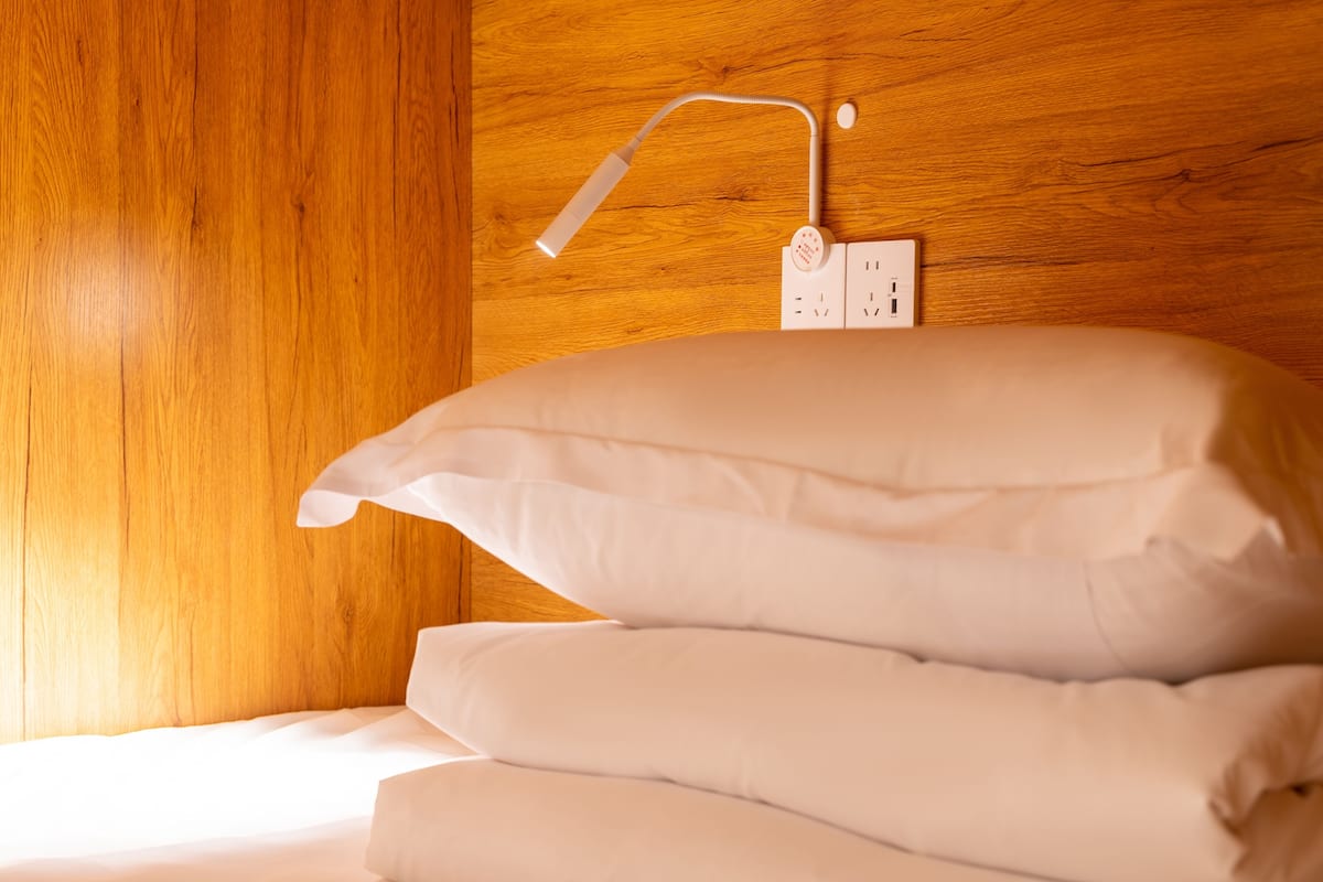 A neatly made bed with white linens, a power outlet, and a wall-mounted lamp on a wooden panel wall.