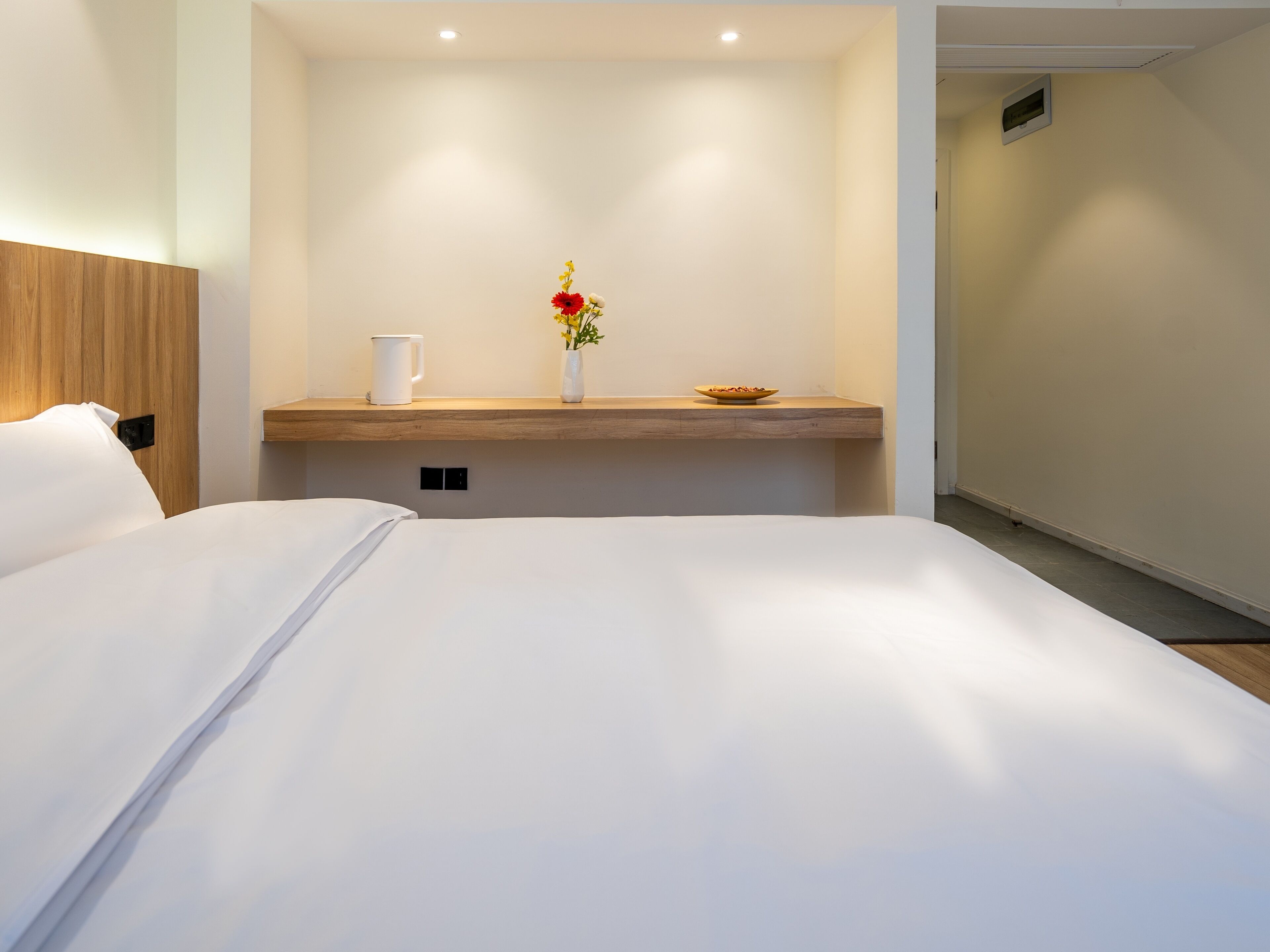 A hotel room with a large bed, a wooden headboard, a bedside table with a white mug and a vase with flowers, and a small bowl.