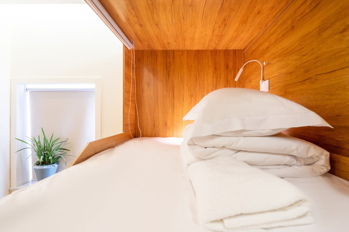 A neatly made bed with white linens and pillows, a wooden headboard, and a small potted plant on the windowsill.