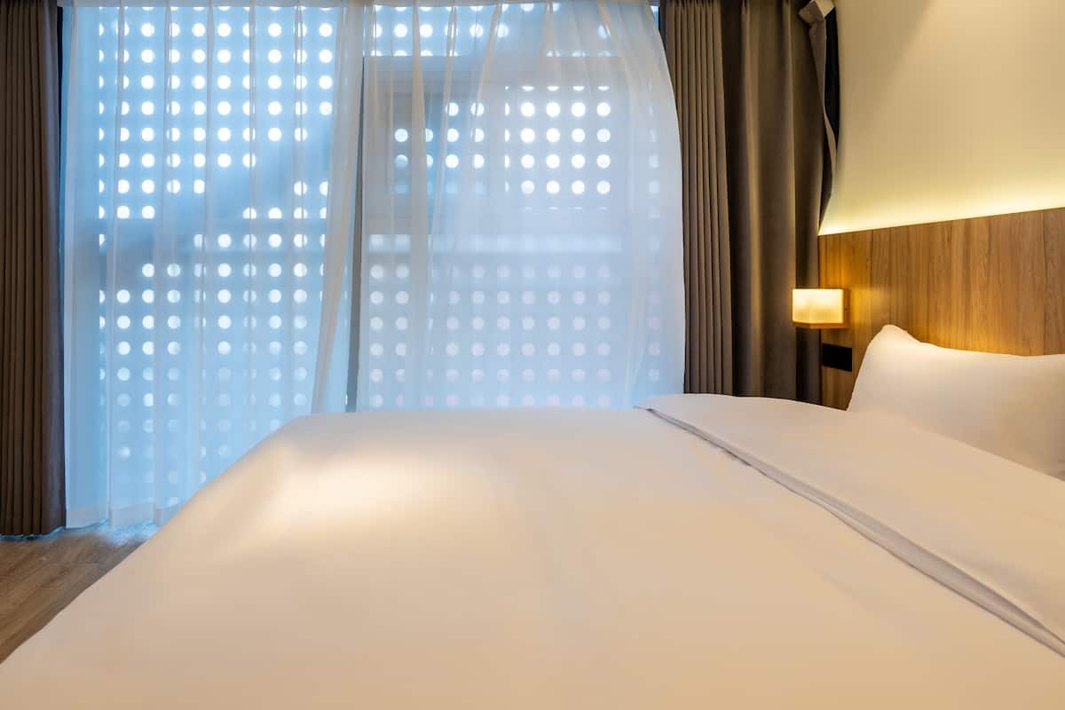 A hotel room with a large bed, a nightstand with a lamp, and a window with sheer curtains.