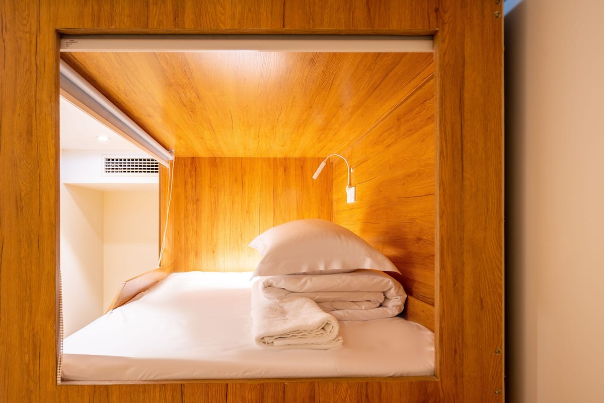 A compact wooden room with a bed, a wall-mounted light, and a visible air vent.