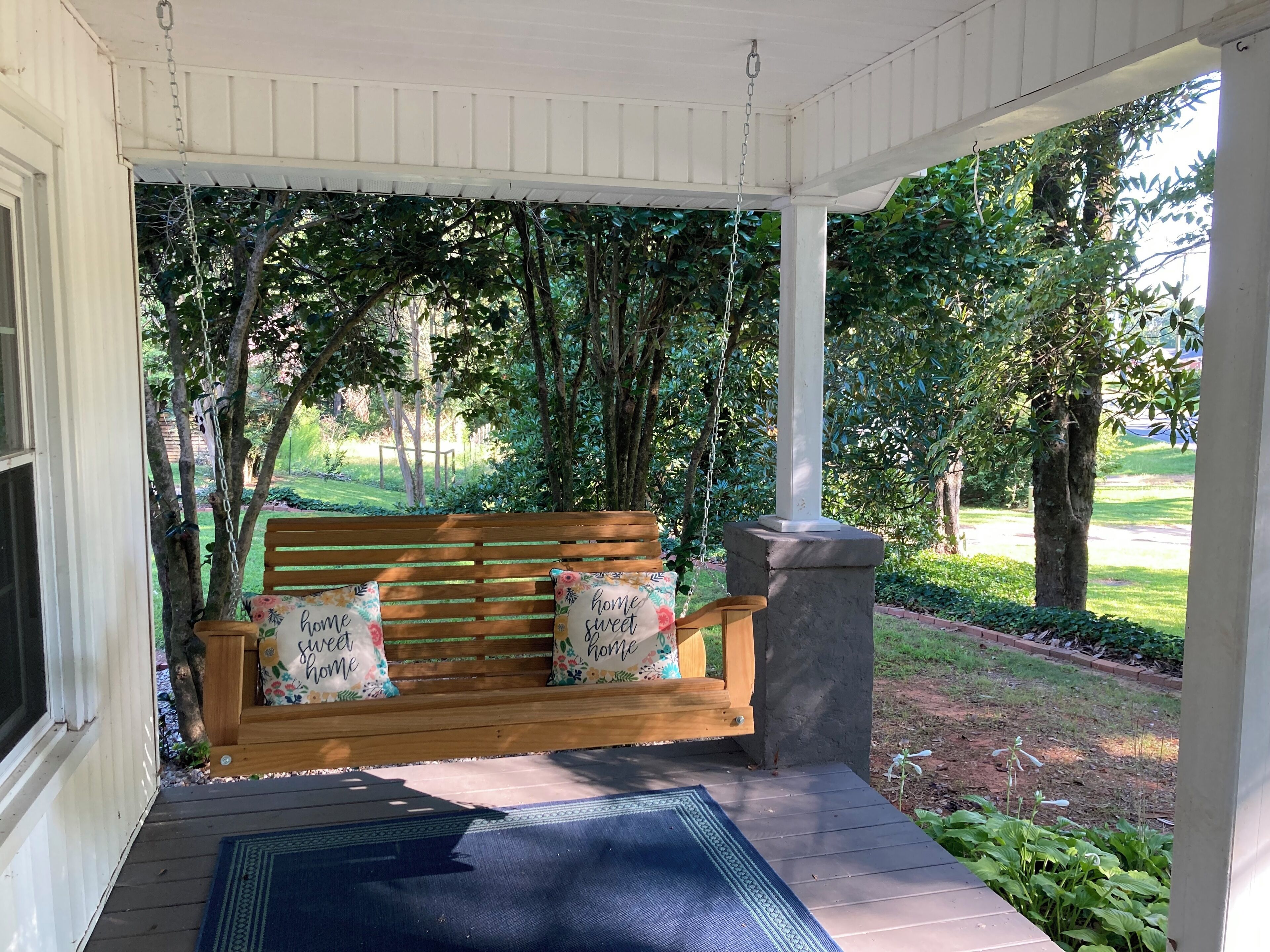 A wooden bench with floral cushions and a stone pillar, set on a porch with a blue mat.