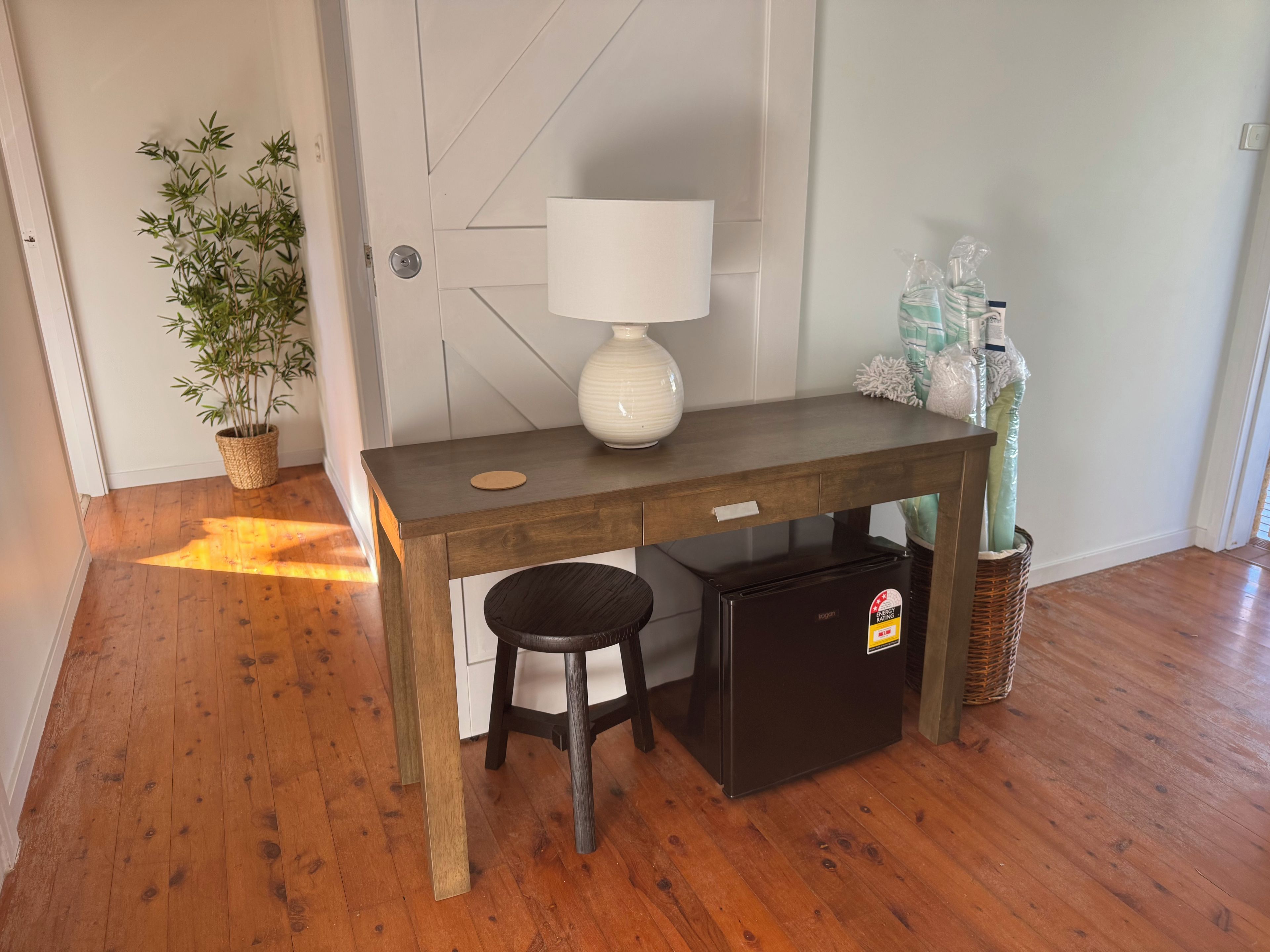 A compact room with a wooden desk, a small stool, a white lamp, and a refrigerator.