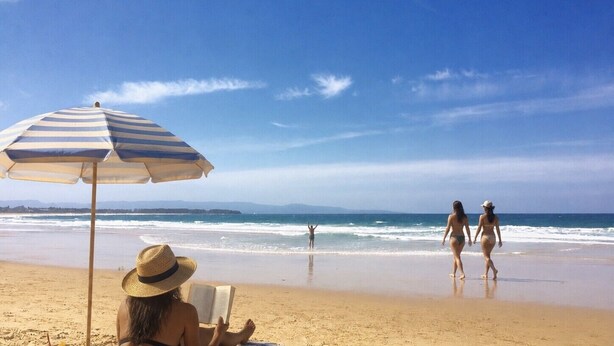 Eine Strandansicht mit einem gestreiften Sonnenschirm, einer Person, die ein Buch liest, und zwei Personen, die am Strand entlanggehen.