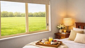 Ein Hotelzimmer mit einem Bett, einem Nachttisch mit Lampe, einem Tablett mit Frühstücksangeboten und einem Fenster mit Blick auf ein grünes Feld.