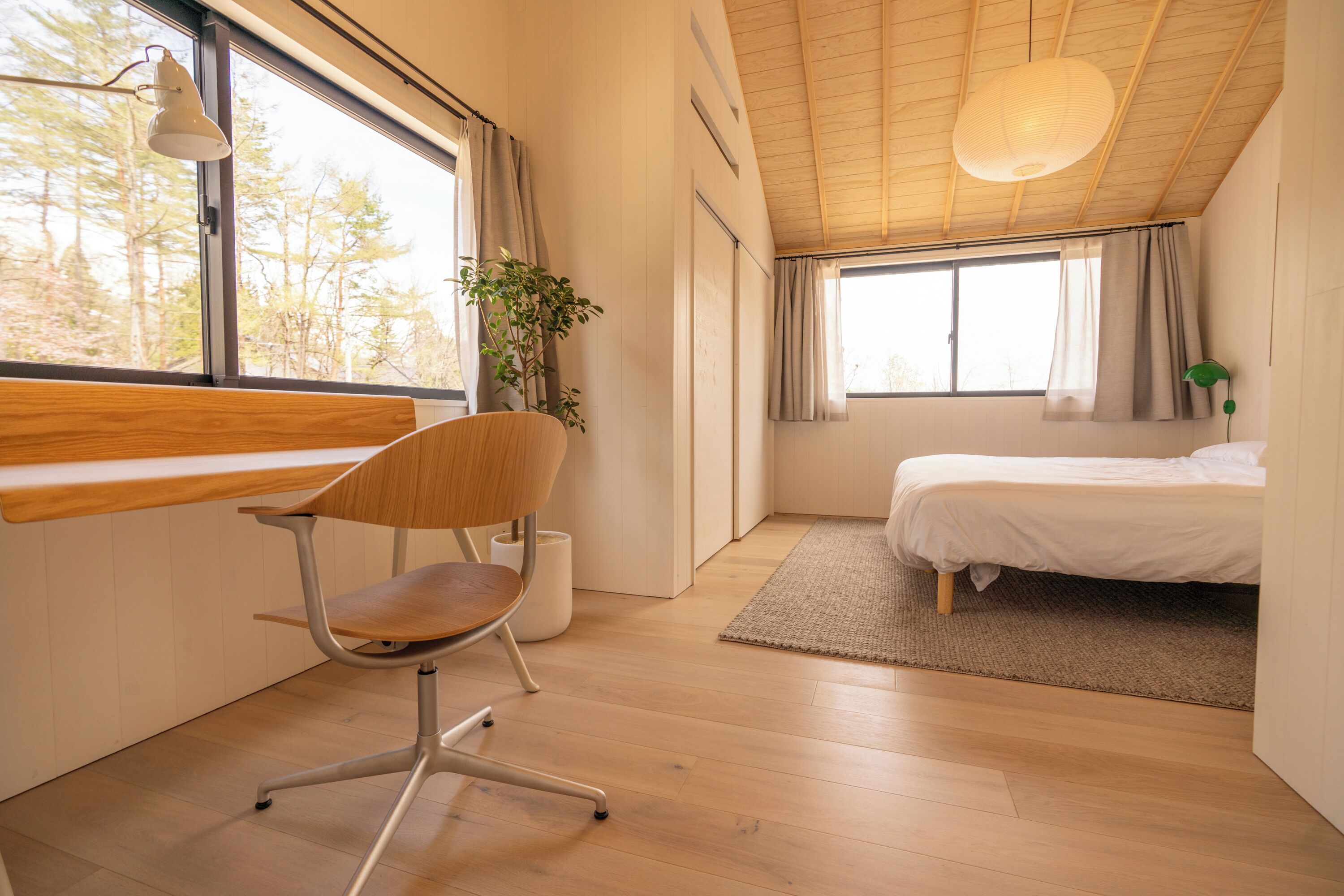Ein modernes Schlafzimmer mit Schreibtisch aus Holz, einem Sessel, einem Bett und einem großen Fenster mit Blick auf Bäume.