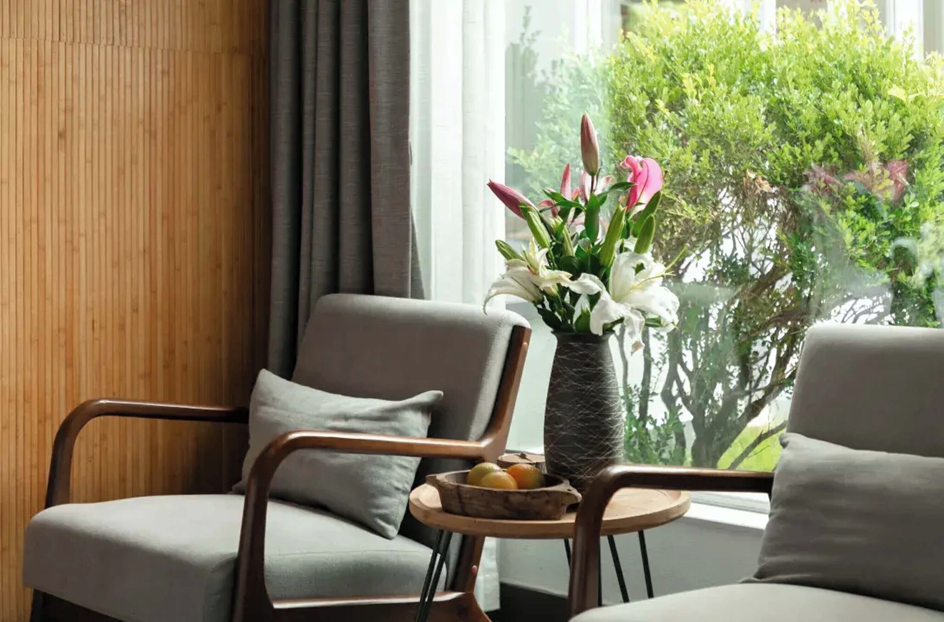 A modern room with two grey armchairs, a small round table with a fruit bowl, and a vase of flowers by a window with a view of greenery.
