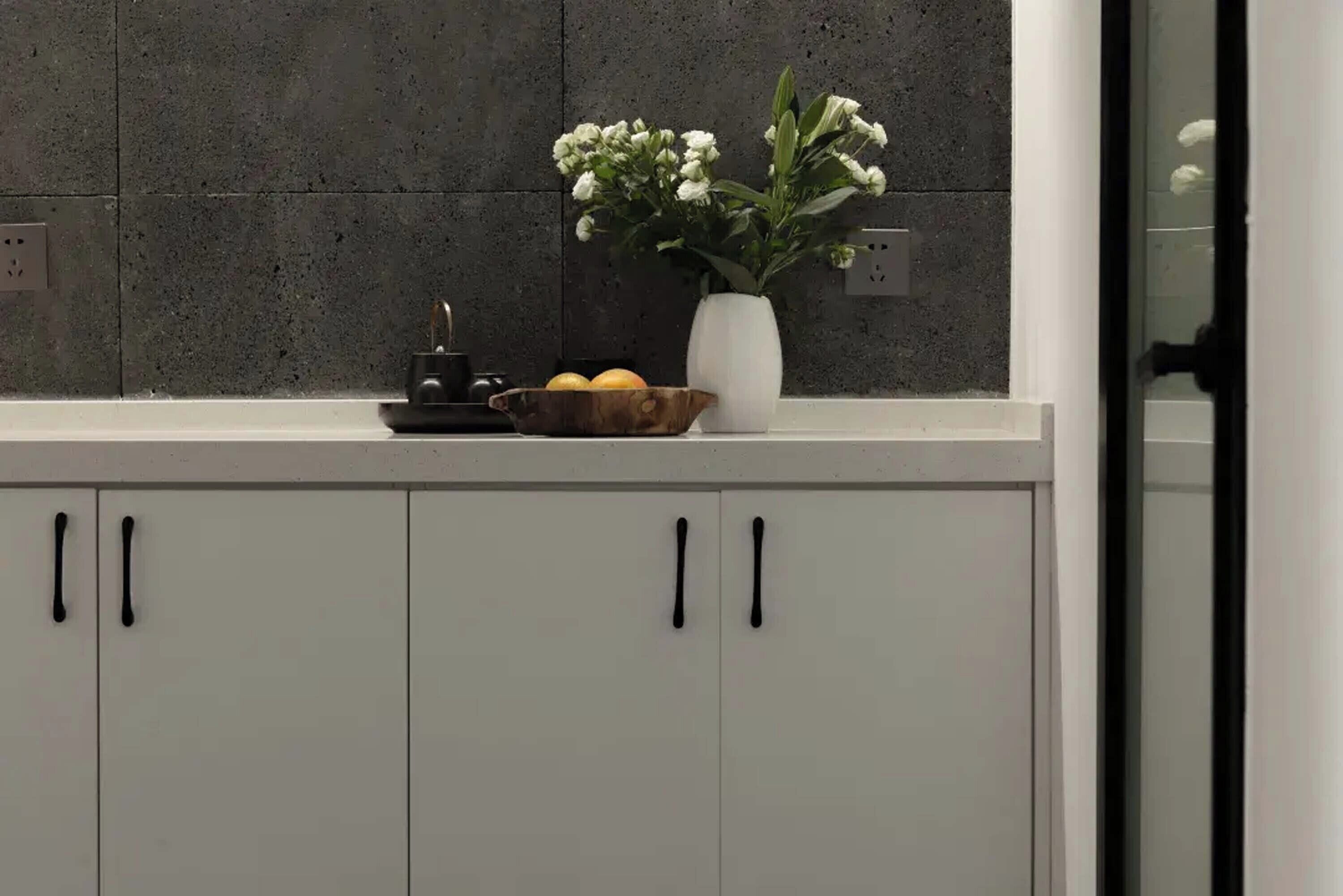 A kitchen counter with a white vase containing white flowers, a wooden bowl with lemons, and a small black appliance.