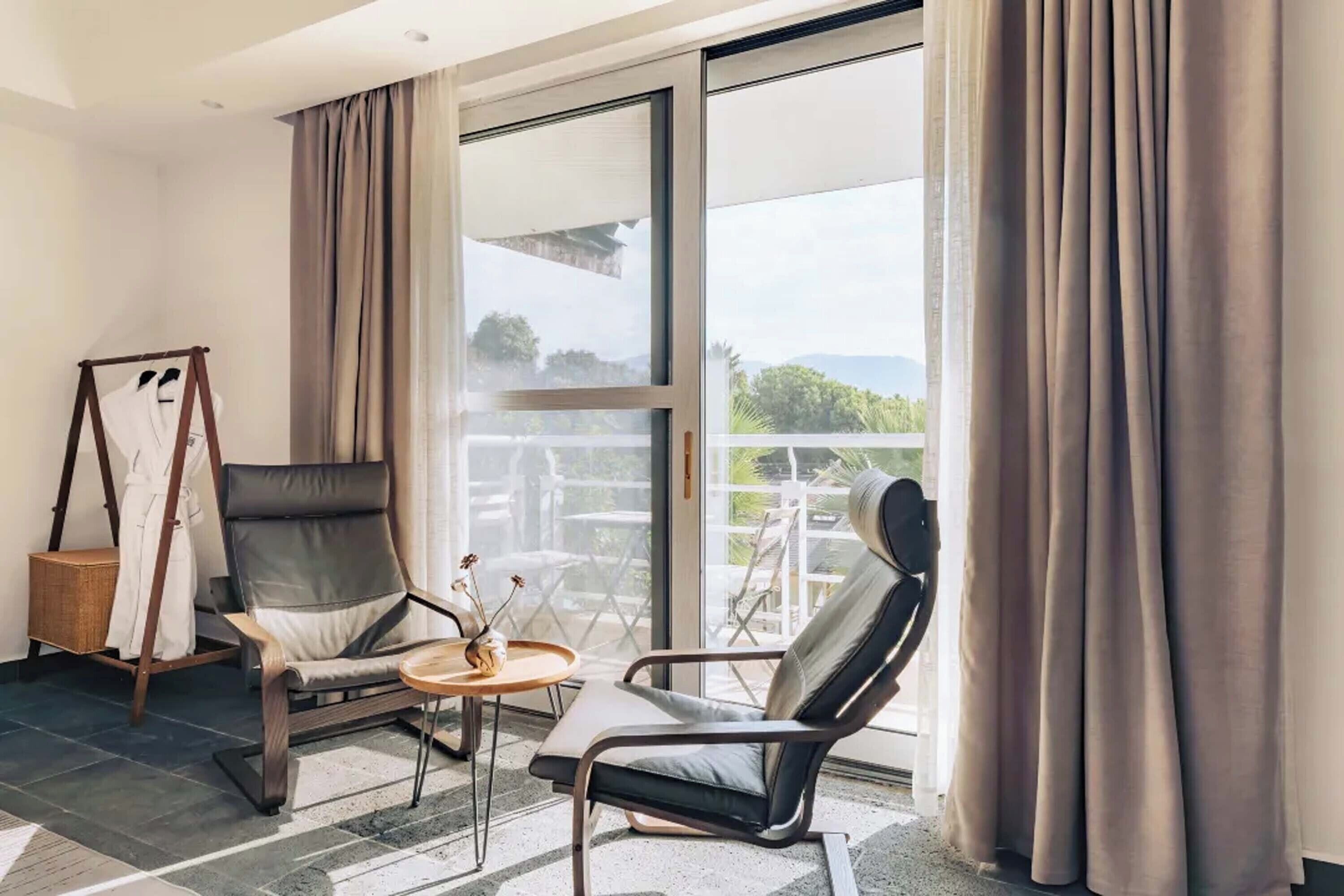 Una habitación moderna con un amplio ventanal corredero que da a un balcón, un sillón de cuero negro, una silla de madera y una pequeña mesa redonda con un adorno.