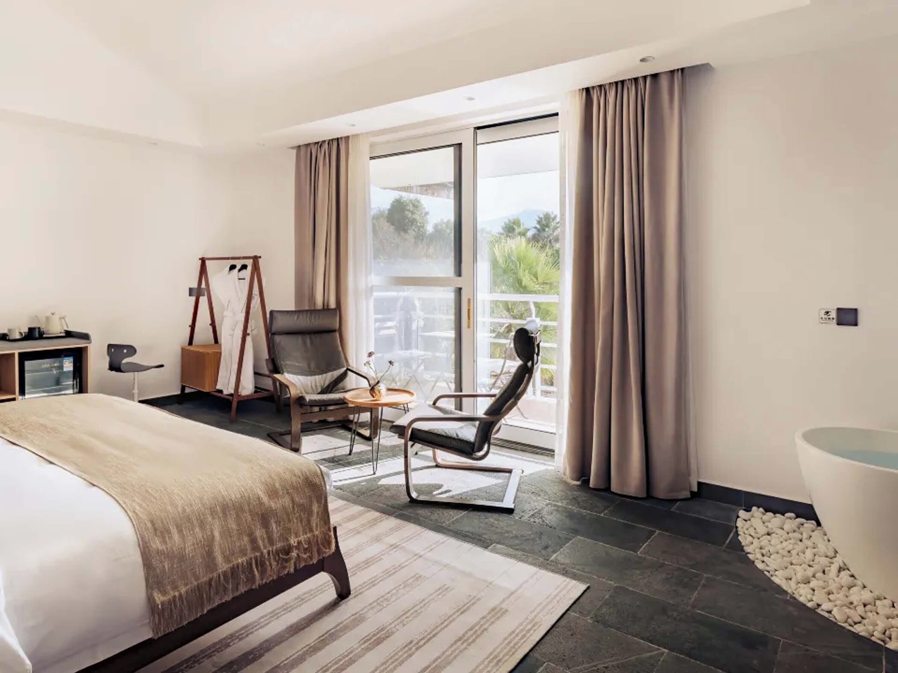 Una habitación de hotel moderna con cama, sillón, escritorio y un ventanal grande.