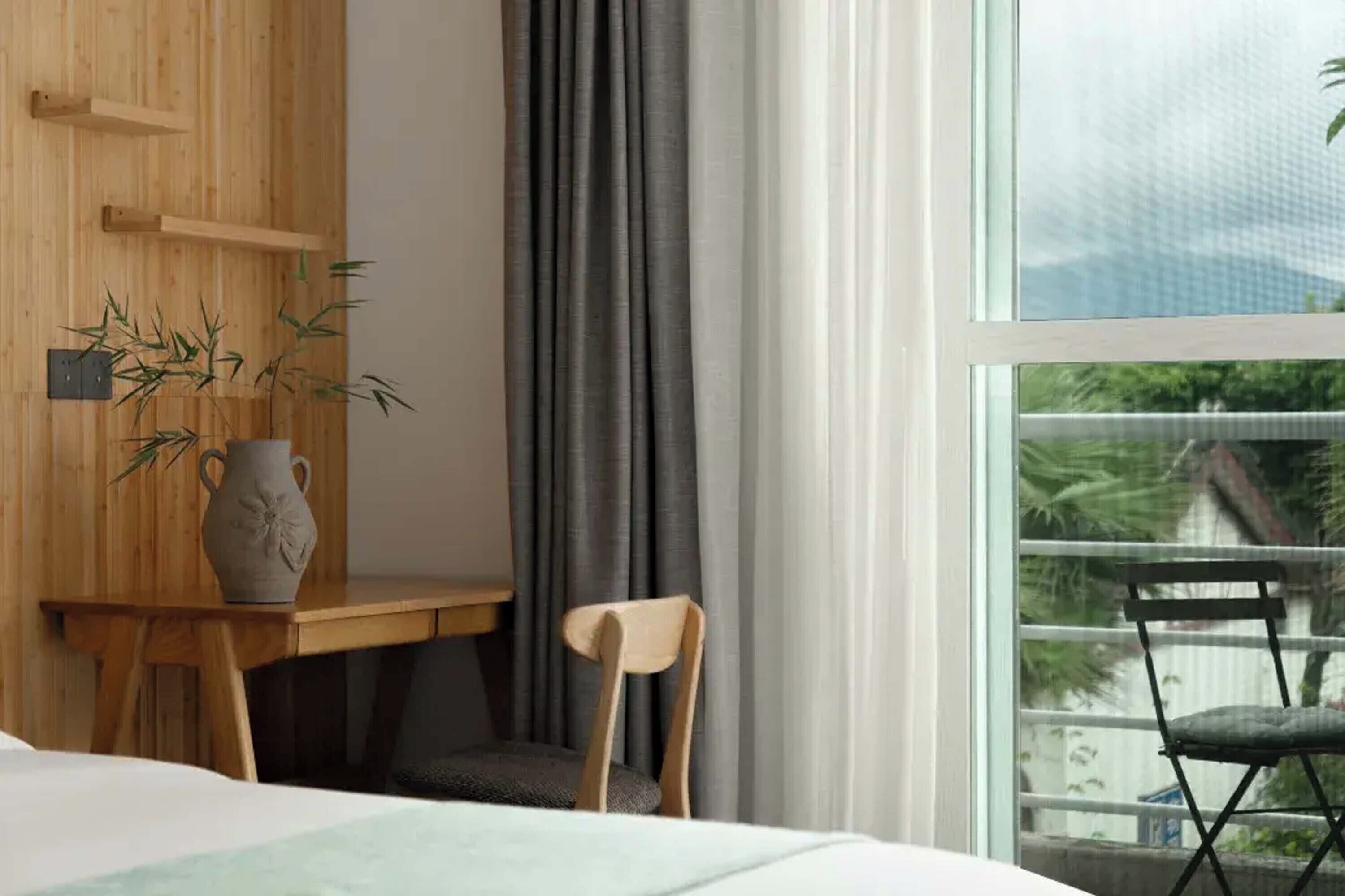 A bedroom with a wooden desk, a chair, a vase with bamboo, and a view of a balcony with a chair.