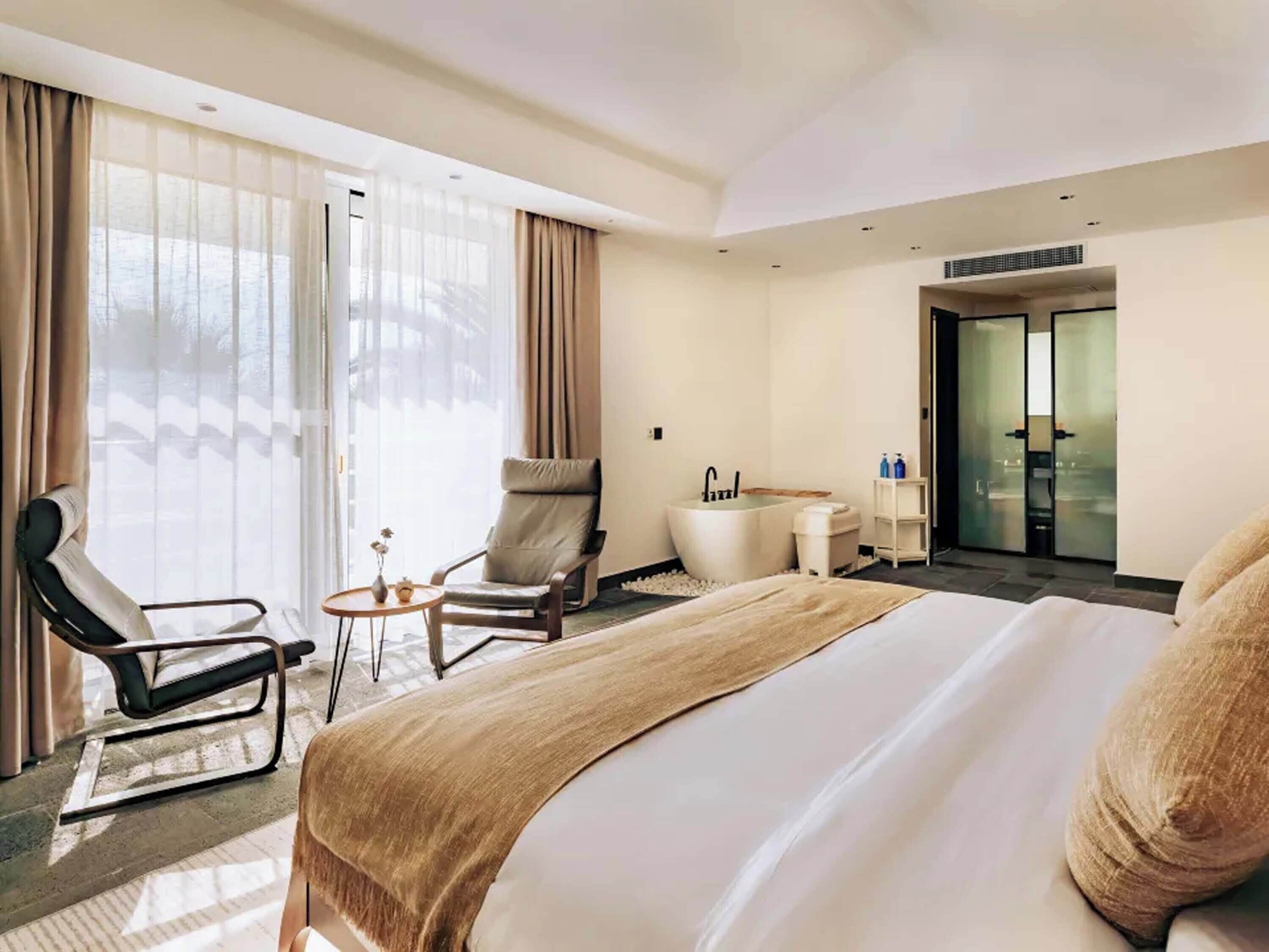 Habitación de hotel con una cama grande, una zona de estar con una silla y una mesita, una bañera y un ventanal con acceso a un balcón.