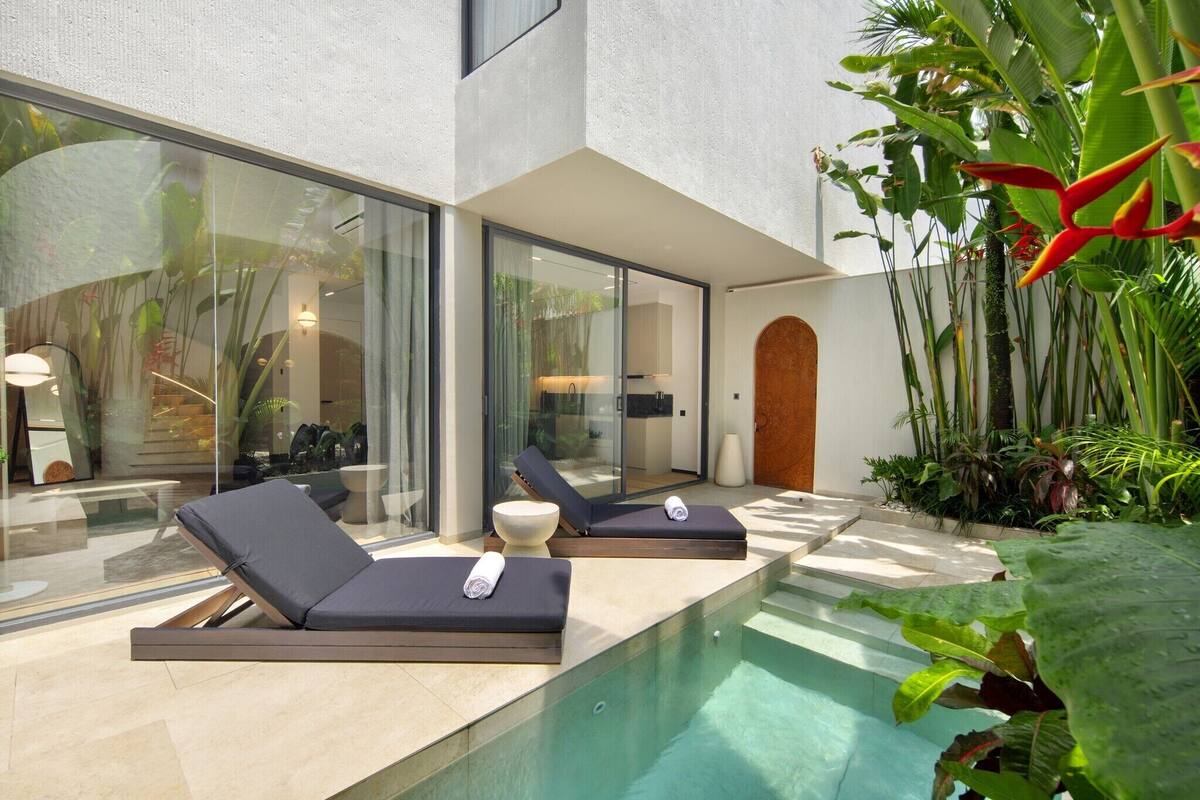 Un área de piscina moderna al aire libre con dos sillones de descanso y una pequeña mesa, rodeada de exuberante vegetación tropical y una pared de vidrio.