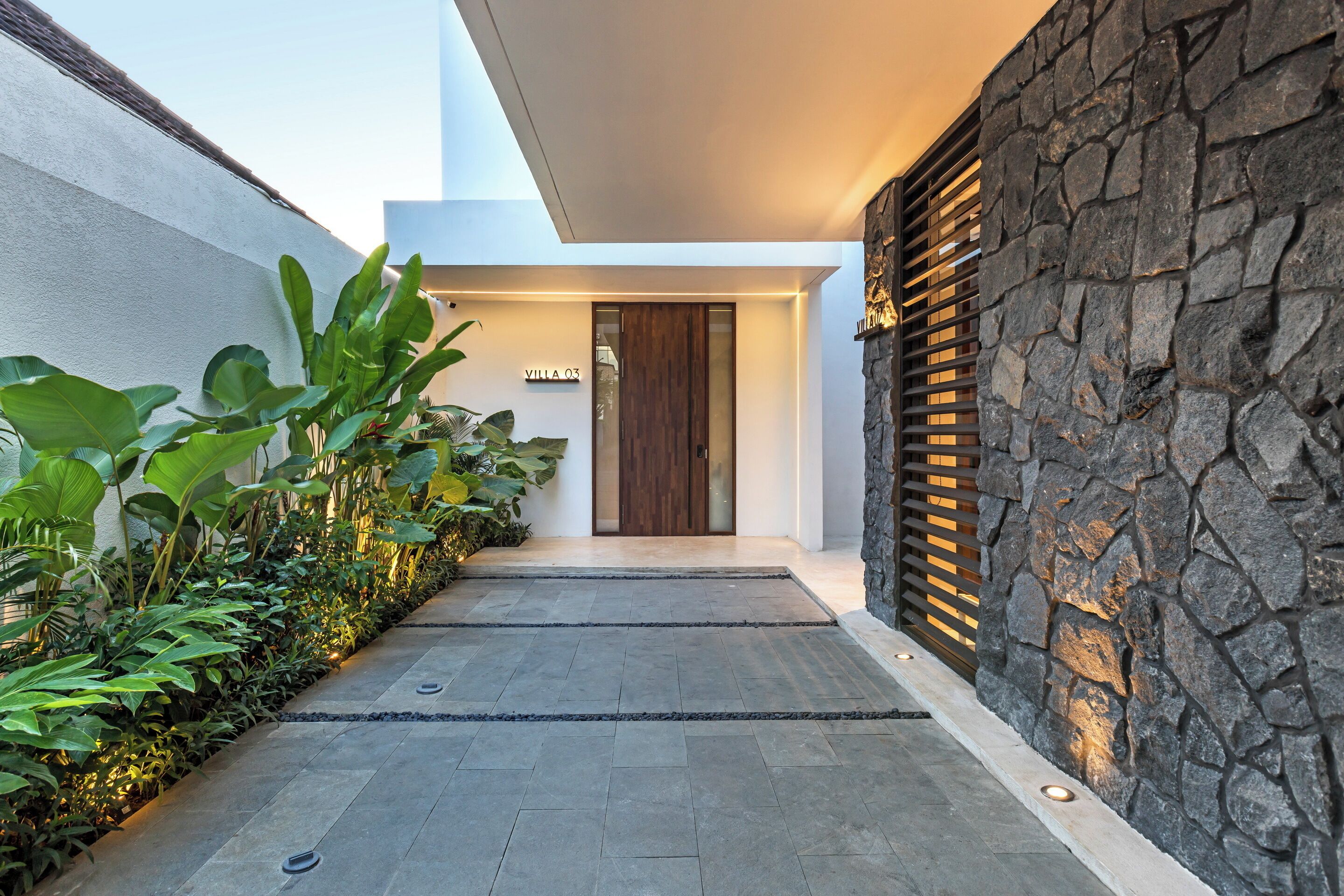 A modern villa entrance with a stone wall, a wooden door, and a pathway leading to the entrance.