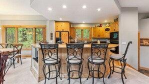 A spacious kitchen with wooden cabinets, a central island with bar stools, and a large window overlooking a forest. - Ski-In Ski out on Skyway • Hot Tub • 4BR • Walk to Town (Breckenridge)