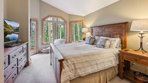 A bedroom with a large bed, a television on a dresser, and a window with a view of trees. - Ski-In Ski out on Skyway • Hot Tub • 4BR • Walk to Town (Breckenridge)