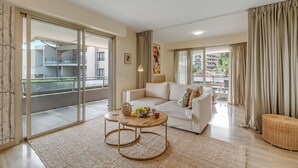 A modern living room with a white sofa, a round wooden coffee table with fruits, and a balcony with a view of buildings and greenery.