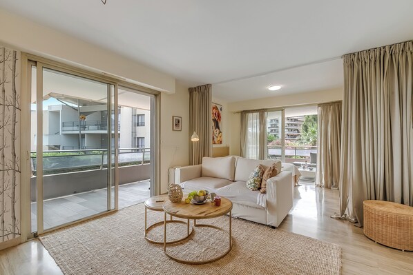 A modern living room with a white sofa, a round wooden coffee table with fruits, and a balcony with a view of buildings and greenery.