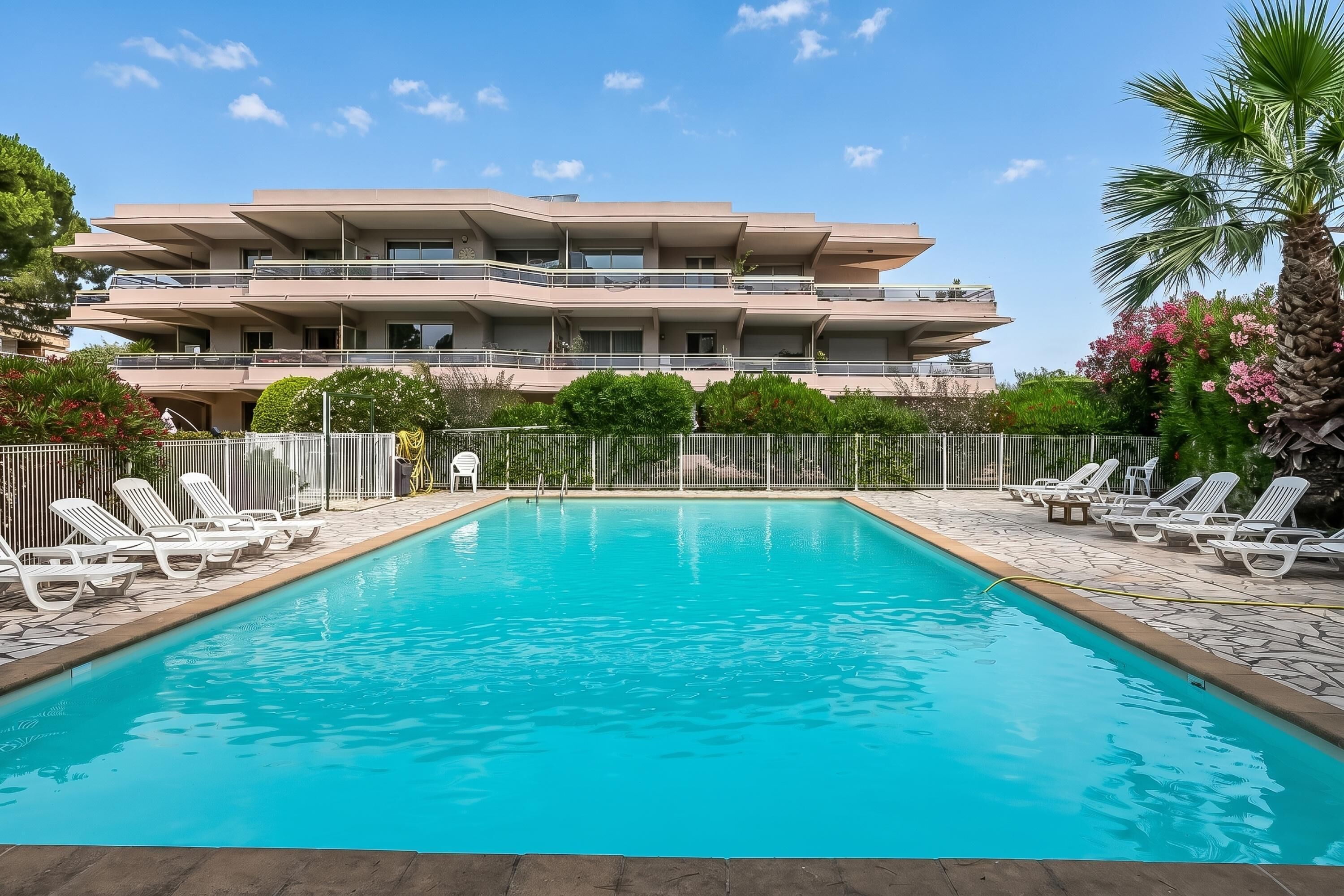 A swimming pool with lounge chairs, surrounded by a fence, and a multi-story building in the background.