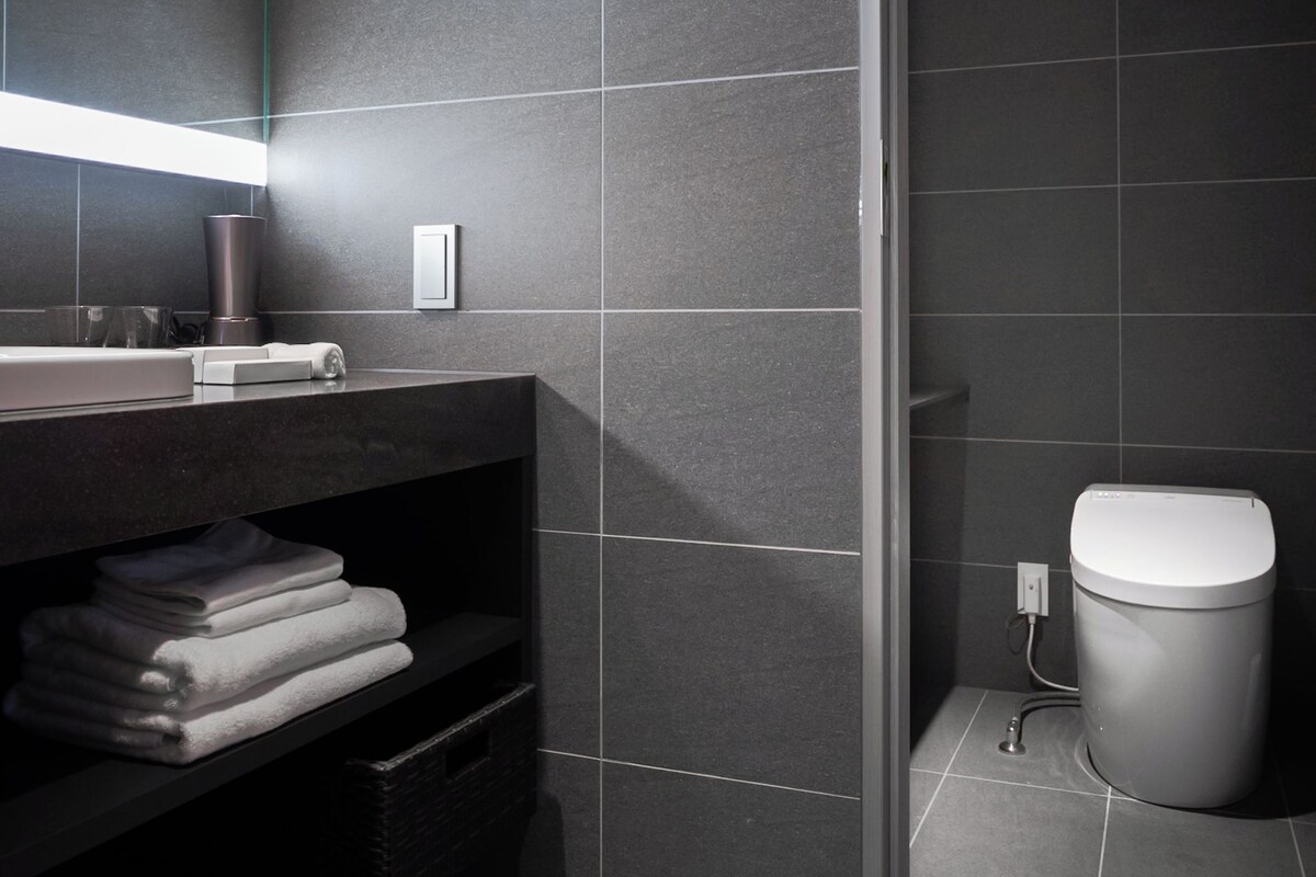 Un bagno moderno con una parete rivestita di piastrelle scure, un water bianco, un lavandino con uno specchio e un portasciugamani.