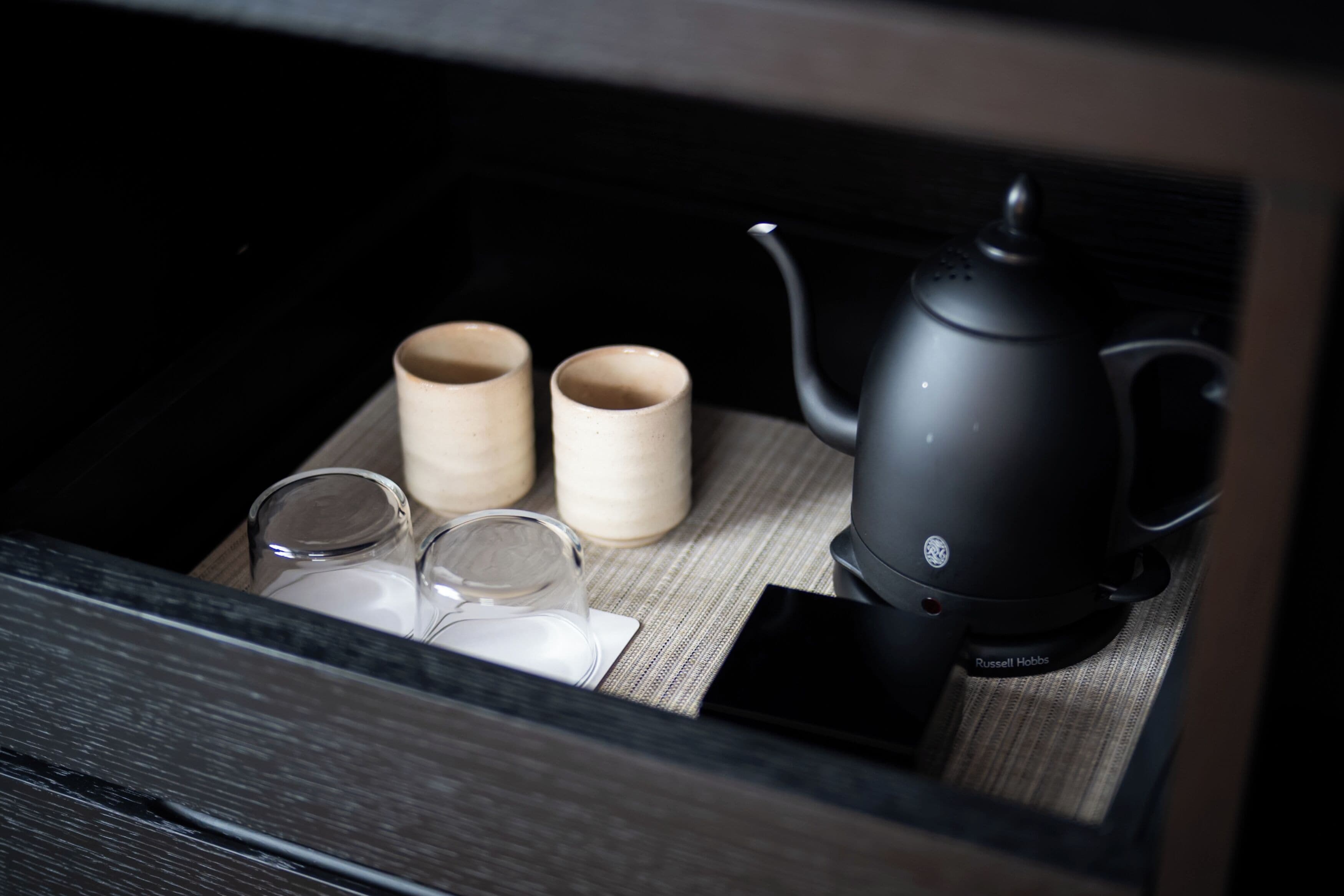 A drawer containing a black teapot, two wooden cups, and two clear glasses on a woven mat.