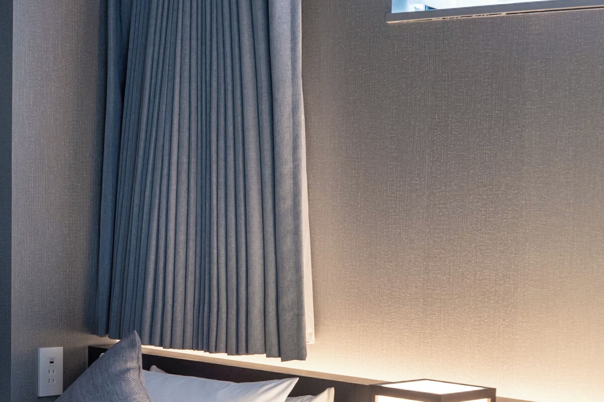 A neatly made bed with white linens and a gray pillow, a bedside table with a lamp, and a window with curtains.