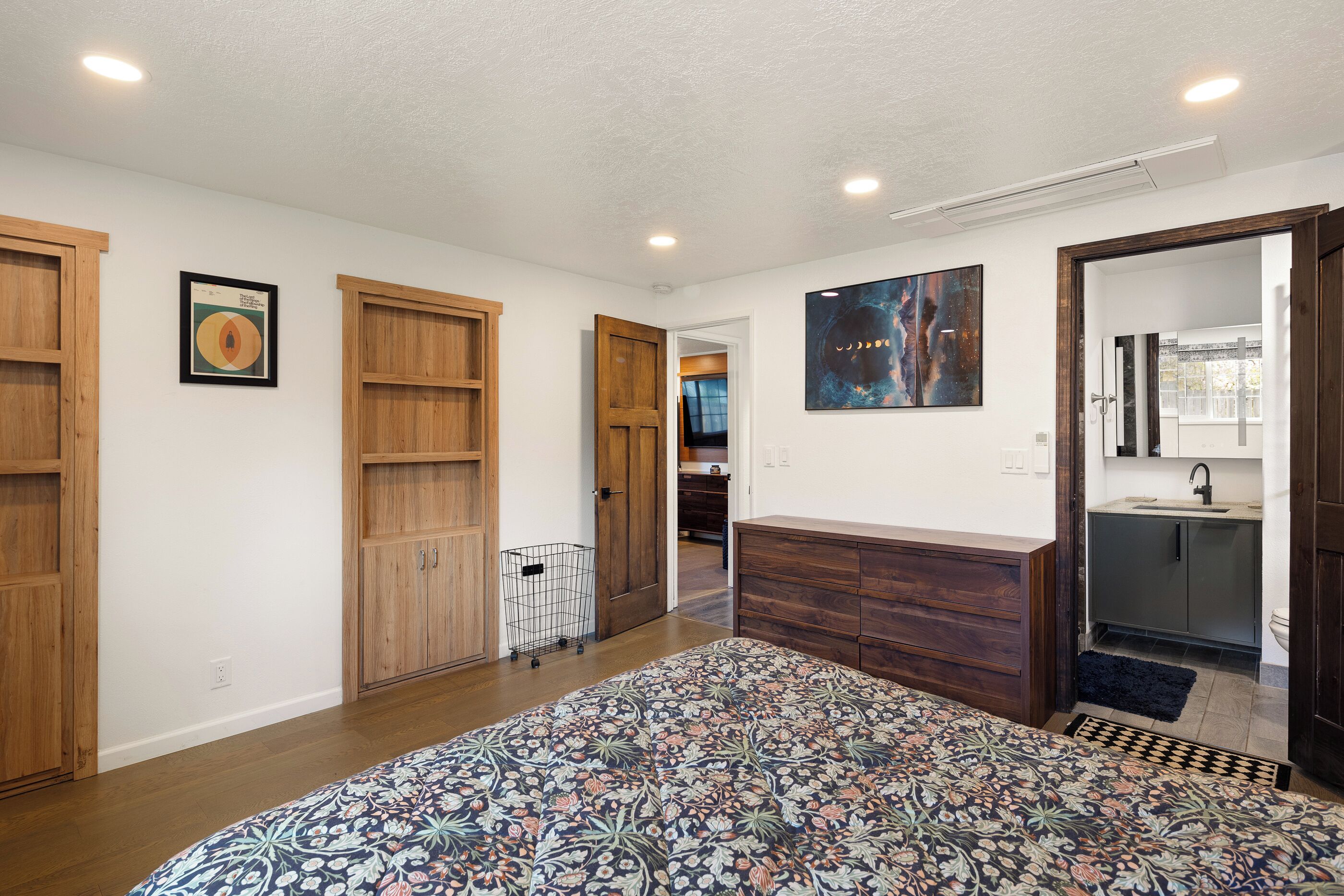 A bedroom with a large bed, wooden wardrobe, a small desk, and a bathroom with a sink and mirror.