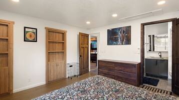 A bedroom with a large bed, wooden wardrobe, a small desk, and a bathroom with a sink and mirror.