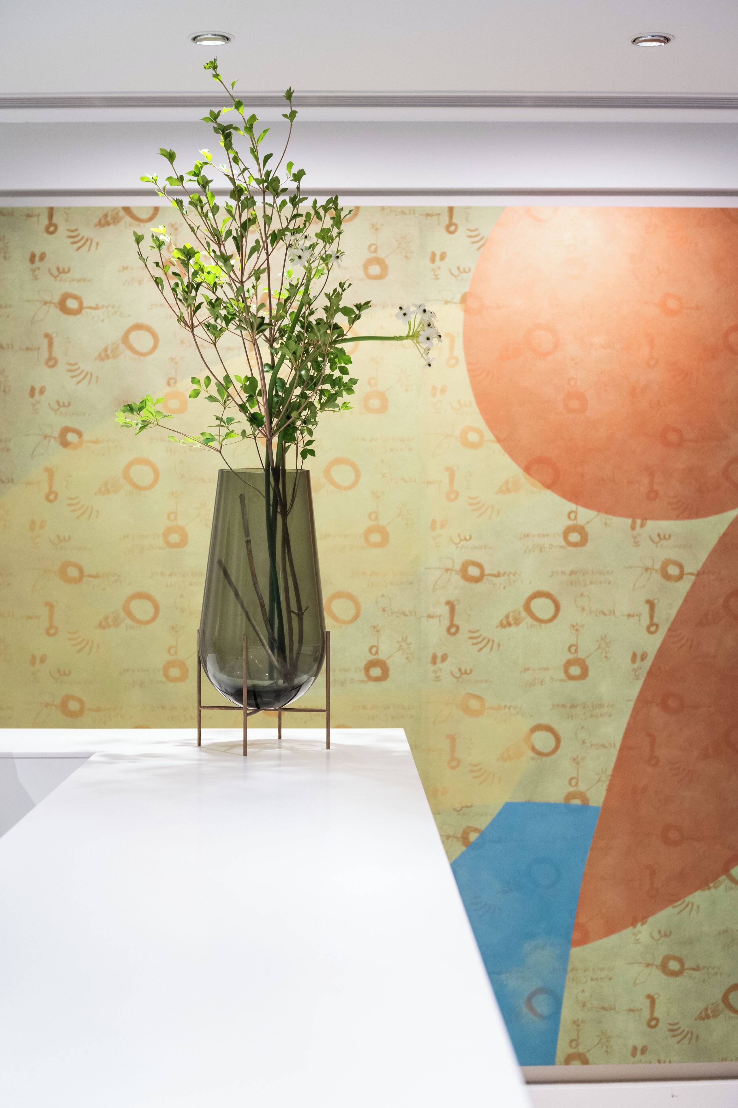 A modern room with a white table, a tall glass vase with green plants, and a wall with abstract patterns.