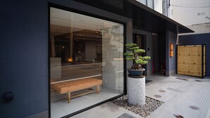 A modern building with a large glass entrance, a wooden bench outside, and a potted bonsai tree. - KIORI Collection Shinmachi (Kyoto)