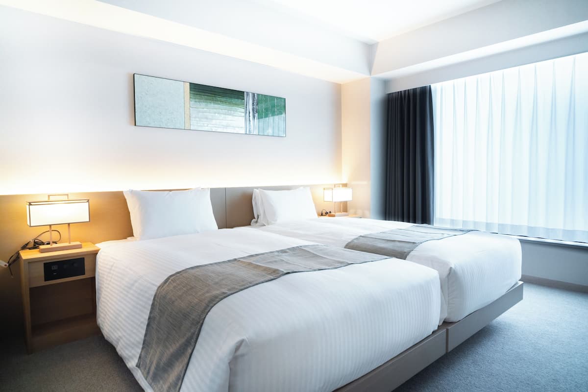A hotel room with two beds, bedside lamps, a nightstand, a wall-mounted artwork, and a large window with curtains.