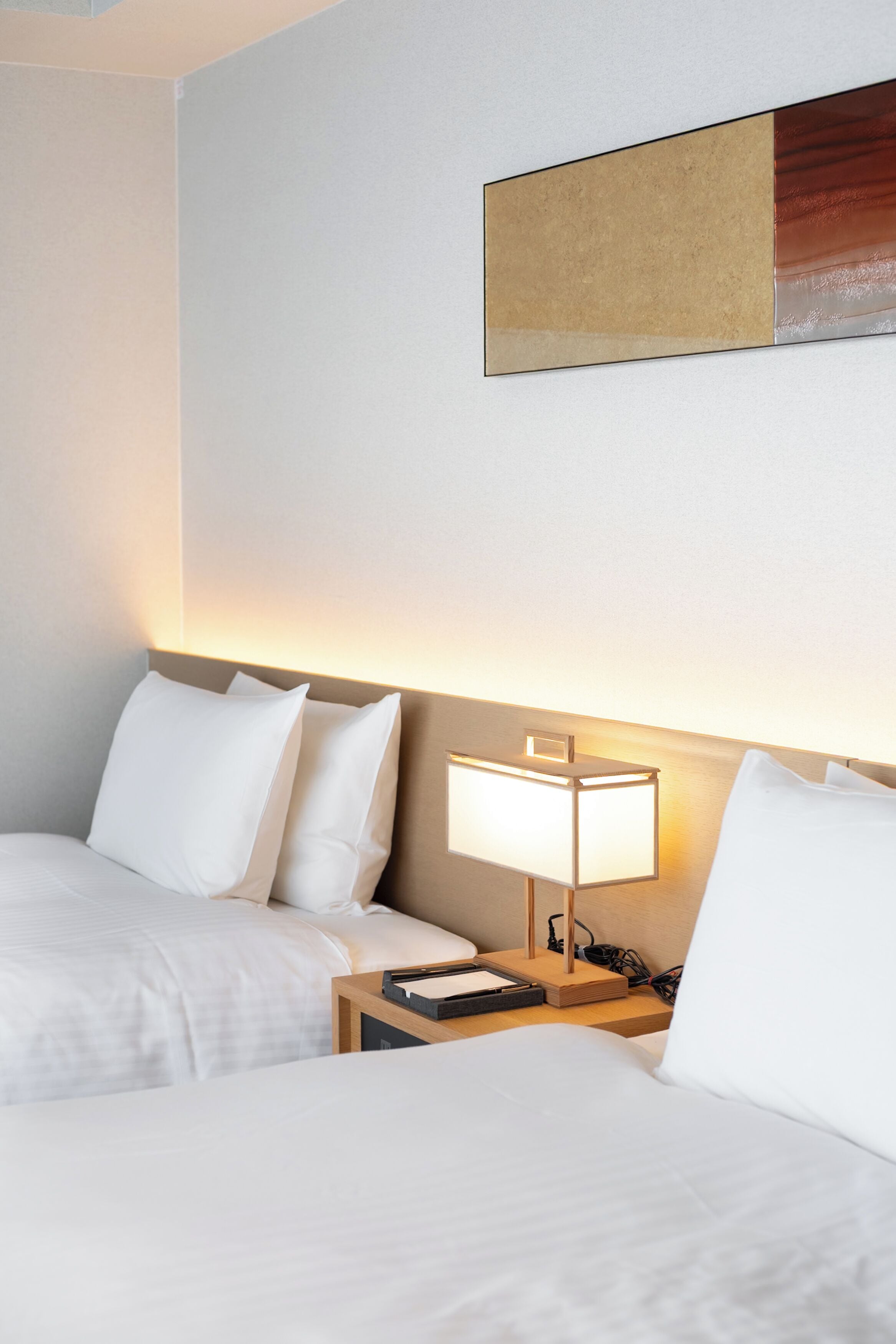 Een hotelkamer met twee bedden, een nachtkastje met een lamp en een kunstwerk aan de muur.