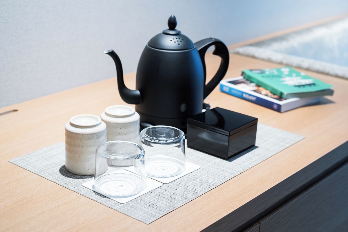 A wooden table with a black teapot, three ceramic cups, two glass cups, and a book.