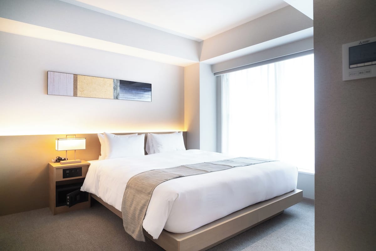 A modern hotel room with a bed, bedside table, lamp, and a large window.