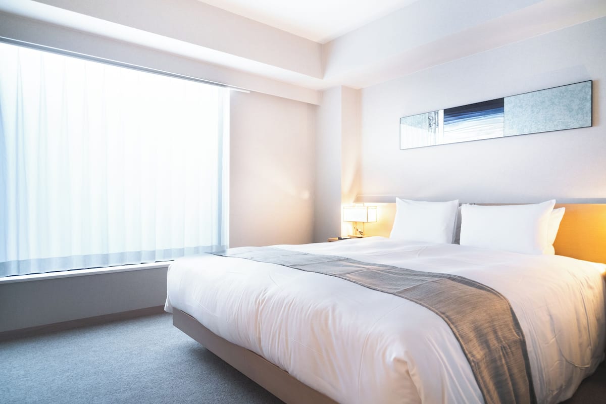 A hotel room with a large bed, a bedside table with a lamp, a window with blinds, and a framed artwork on the wall.