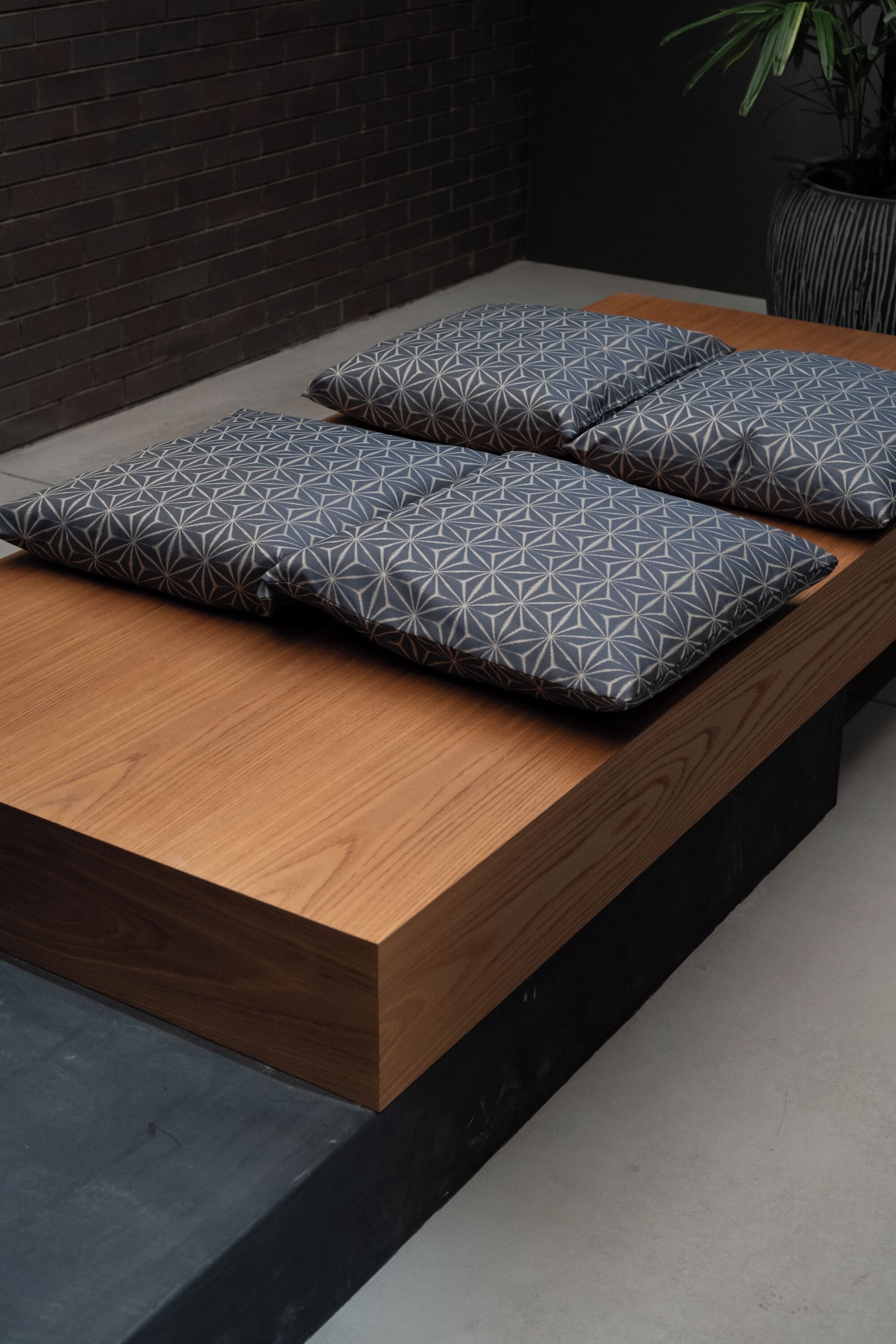 Ein modernes Bett mit geometrisch gemusterten Kissen, einem Holzkopfende und einer dunklen Ziegelwand.