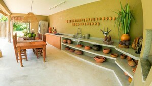 A kitchen with a dining area, featuring a wooden table and chairs, a refrigerator, and shelves with pots and decorative items.