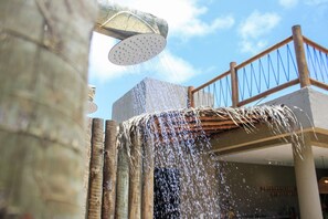 A showerhead with water flowing over a wooden structure, a balcony with a railing, and a building in the background. - MHV Milagres - Casa Aruã (Passo de Camaragibe)