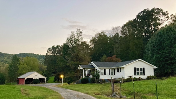 A rural house with a gravel driveway leading to a front porch, surrounded by greenery and hills.