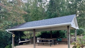 A covered outdoor seating area with a picnic table and benches, surrounded by greenery.