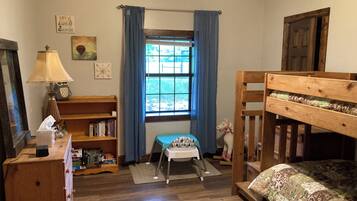 Ein gemütliches Schlafzimmer mit einem Holzbett, einem kleinen Schreibtisch mit Lampe, einem Bücherregal und einem Fenster mit blauen Vorhängen.