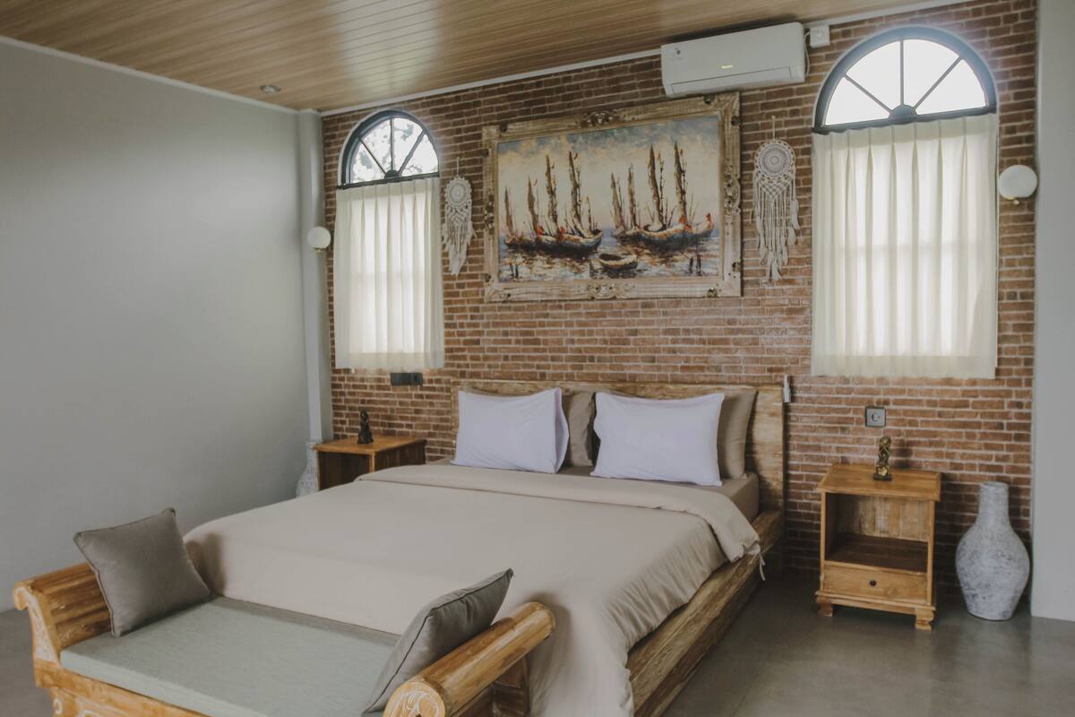 A bedroom with a large bed, two nightstands, a wall-mounted artwork, and a brick wall.