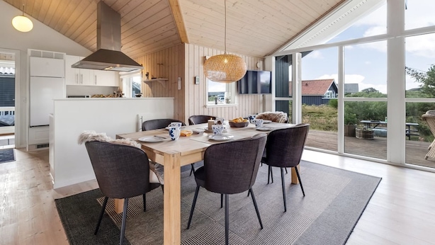 Une cuisine moderne avec des poutres en bois, un coin repas doté d’une grande table et de chaises, et une vue sur l’extérieur grâce à de grandes fenêtres.