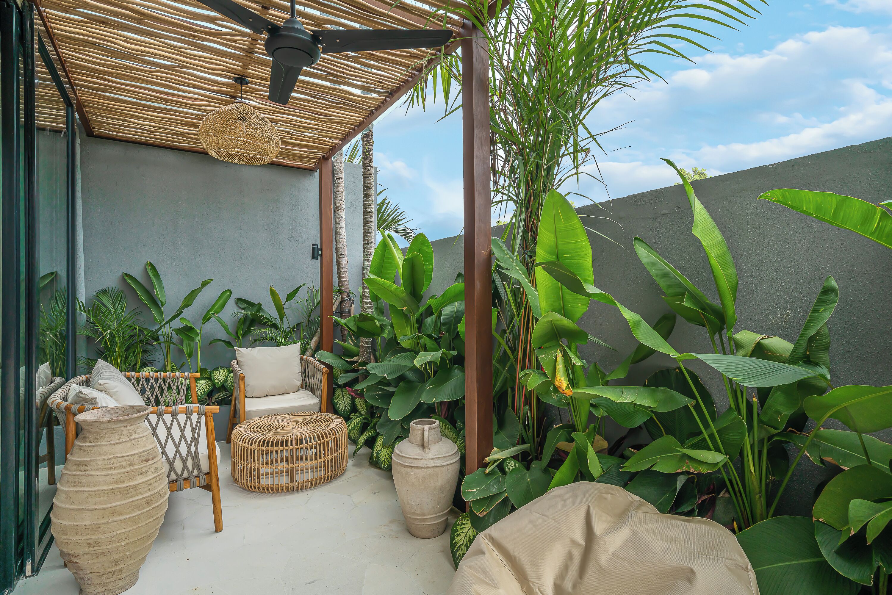 A covered outdoor seating area with wicker furniture, large potted plants, and a ceiling fan.