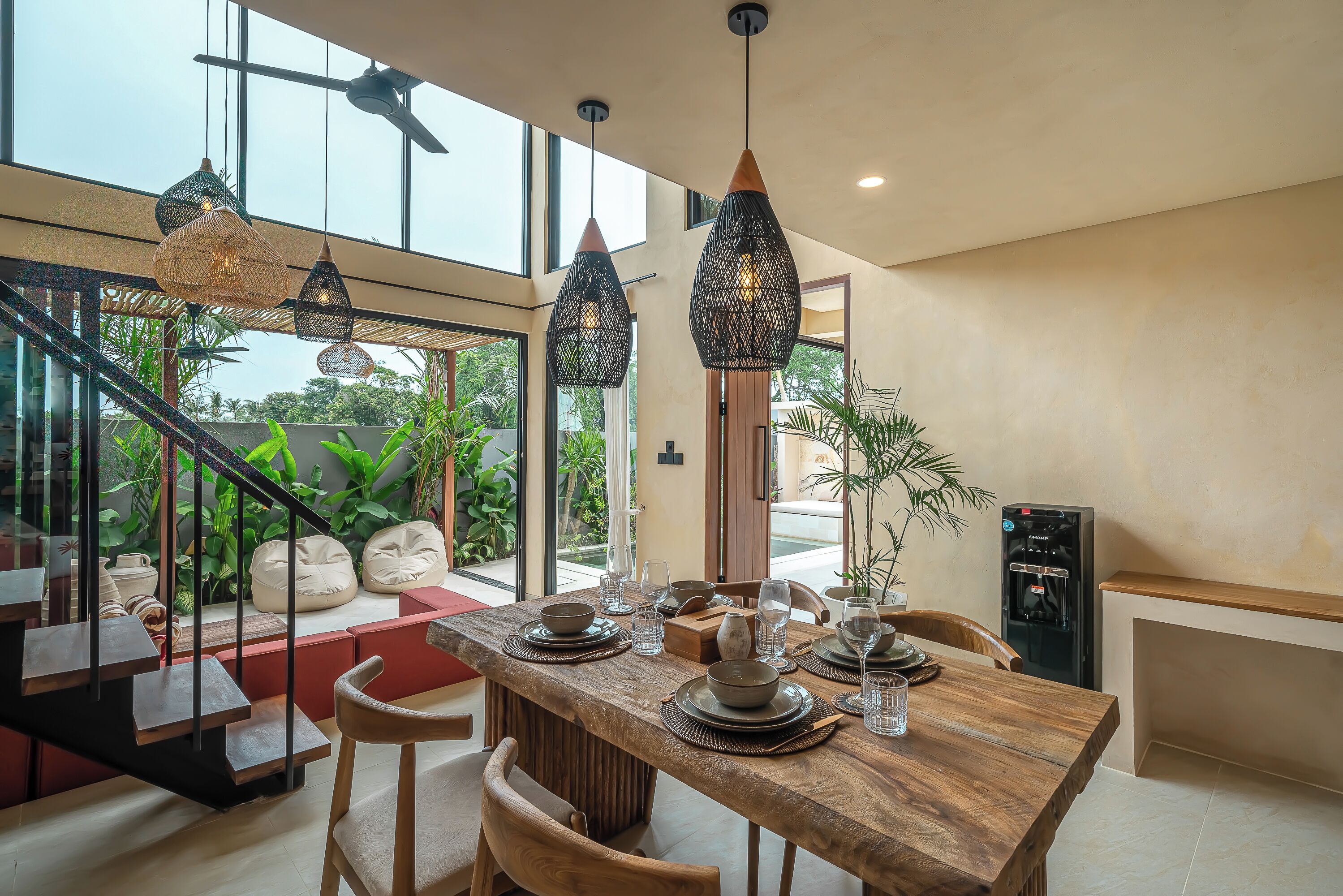 A modern dining area with a wooden table set for four, surrounded by large windows and a staircase.