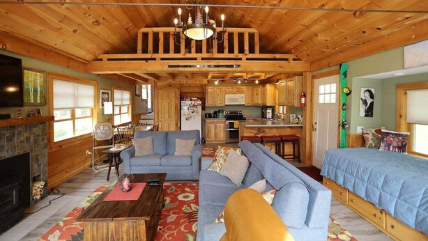 Interior rústico de una cabaña con sala de estar, sofá, chimenea, cocina y un dormitorio en lo alto.