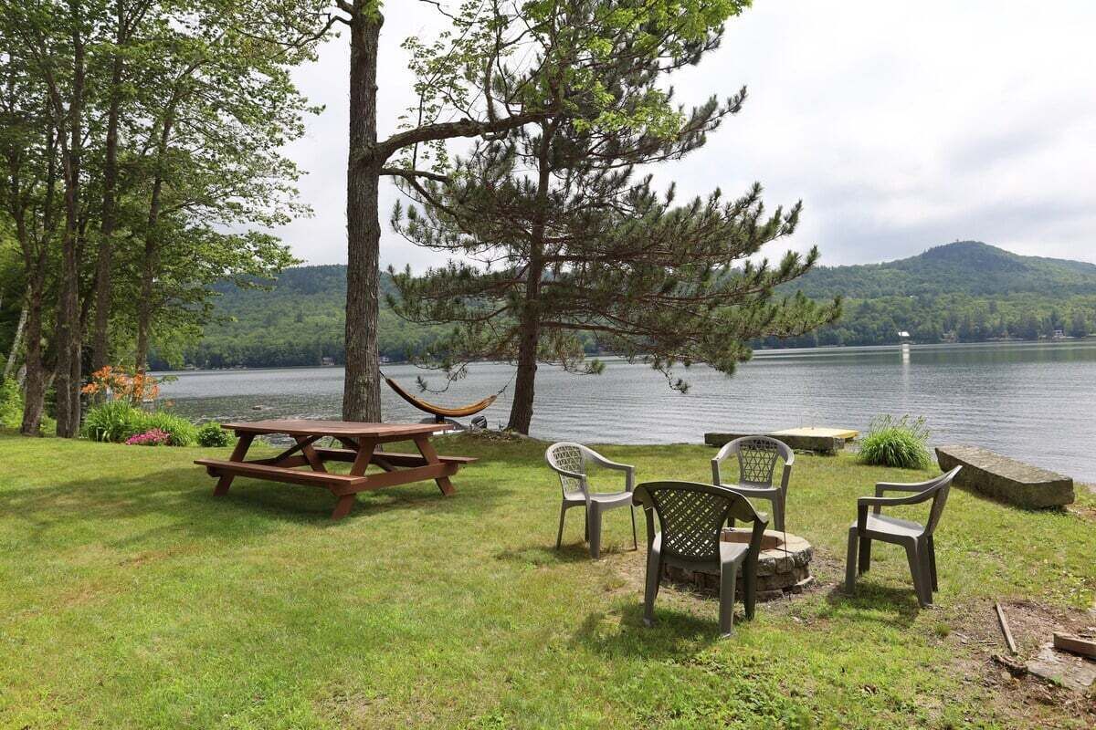 A picnic area with a wooden table and chairs, a hammock, and a view of a lake with trees and hills in the background.