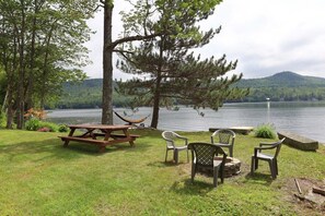 A picnic area with a wooden table and chairs, a hammock, and a view of a lake with trees and hills in the background.
