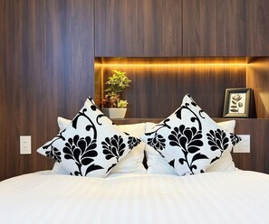 A hotel room with a wooden headboard, a bedside plant, a framed picture, and a bed with white bedding and black floral pillows. - R34A9CC_903 / Koto-ku Tokyo (Koto-ku)
