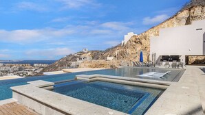 A infinity pool with clear blue water, a white building, and a view of the coastline. - Oceanview Pedregal Condo: Jacuzzi, Balcony & (Cabo San Lucas)
